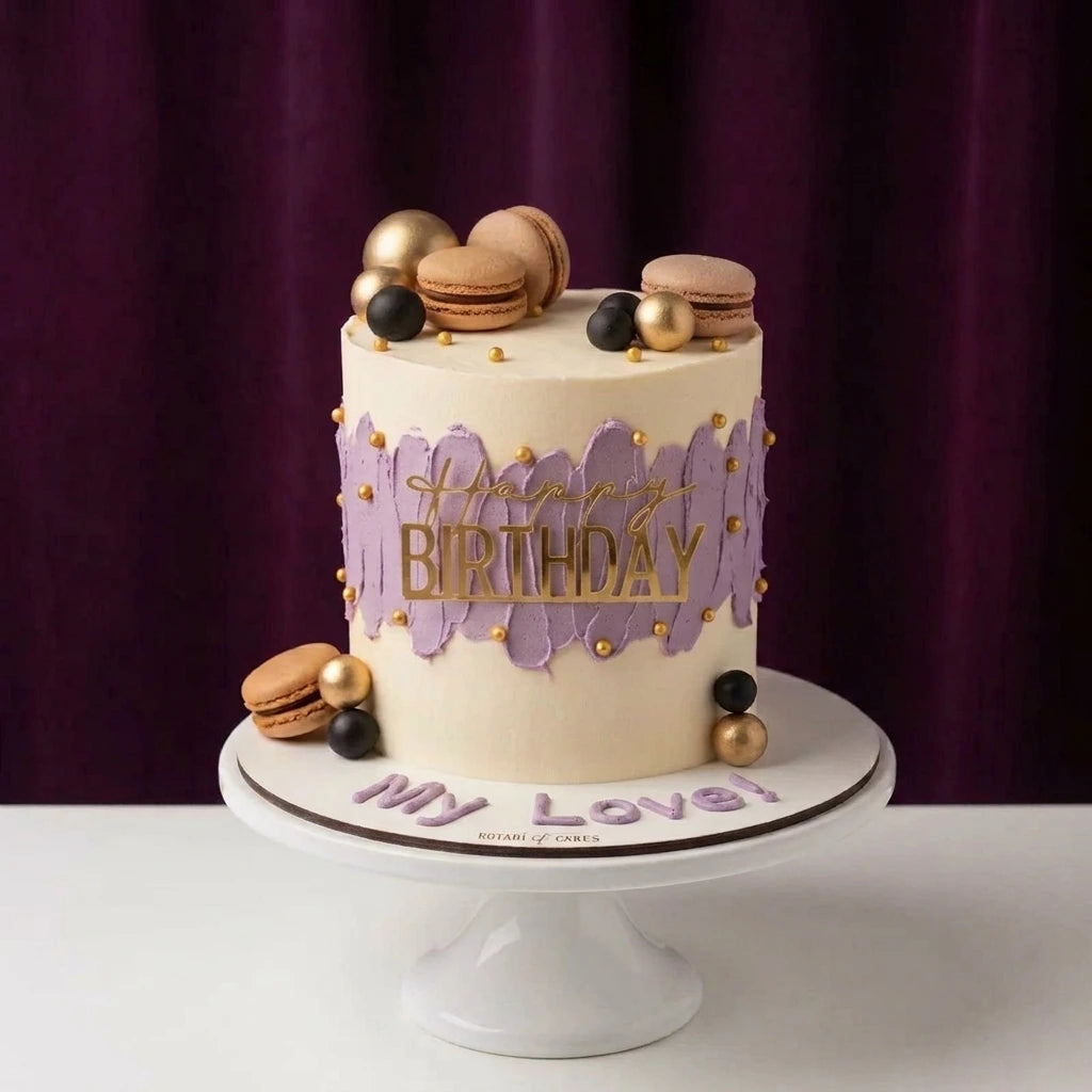 A frosted birthday cake on a white pedestal stand, decorated with purple brushstroke patterns, gold lettering, macarons, and gold and black spherical accents, set against a dark purple curtain background.