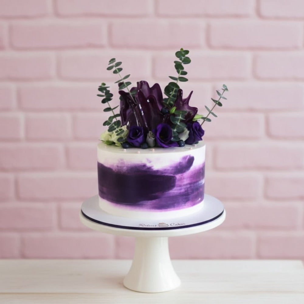 Cake with purple and white design on a white stand against a pink brick wall.