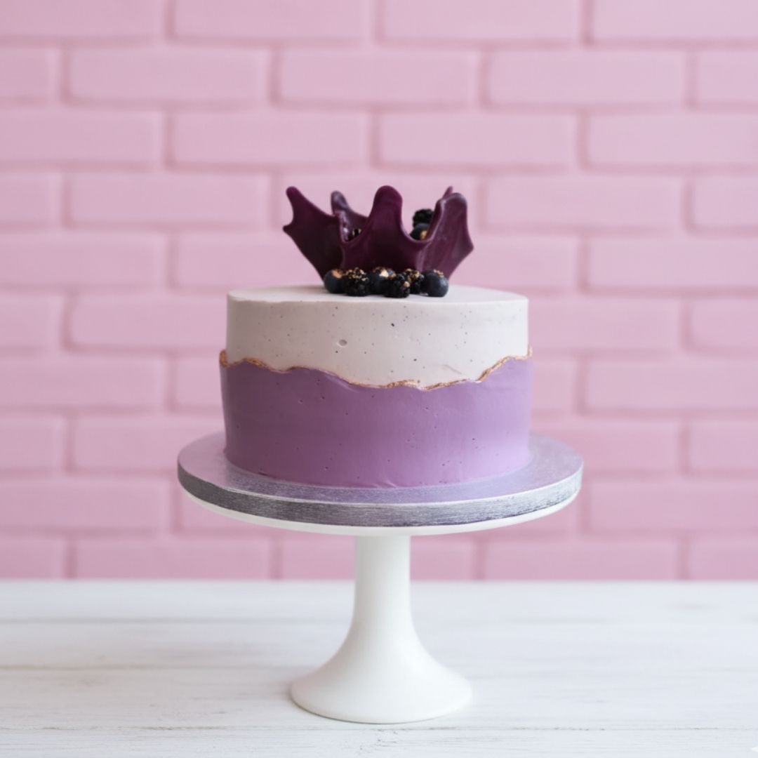 purple cake with purple and white frosting on a white stand against a pink brick wall.