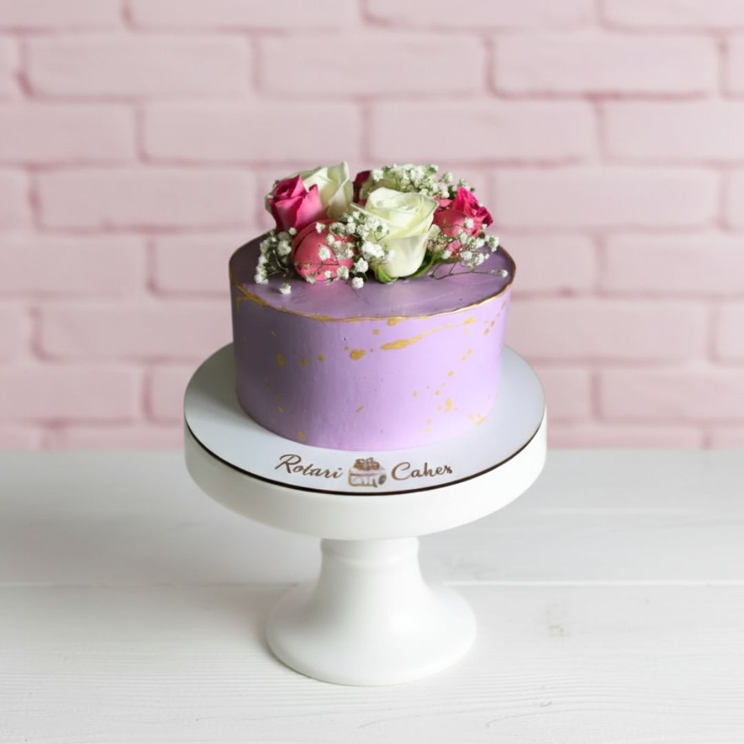 Purple cake with floral decoration on a white stand against a pink brick wall.