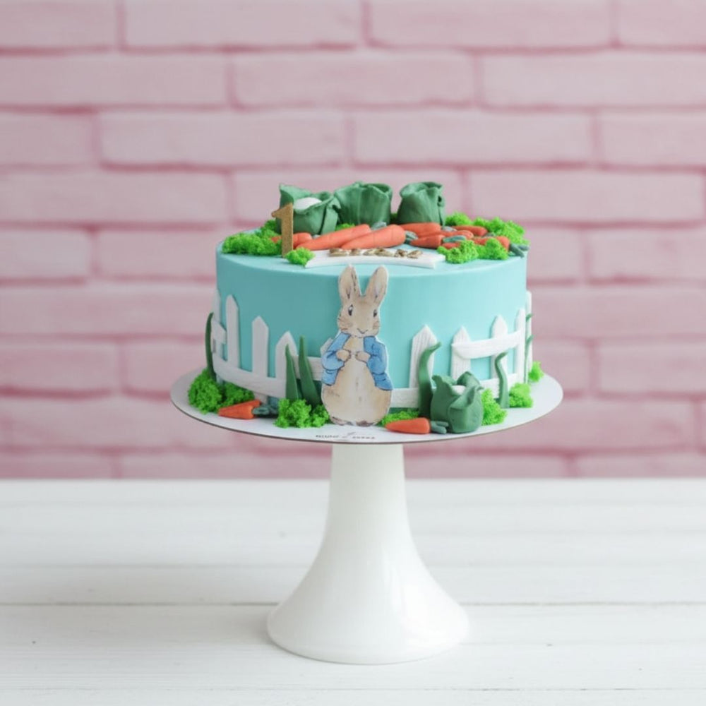Cake with a rabbit design and garden elements on a white stand against a pink brick wall.