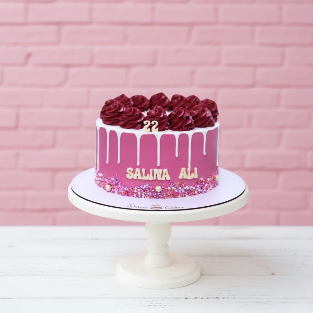 Pink cake with red roses and '22' on a white stand against a pink brick wall.
