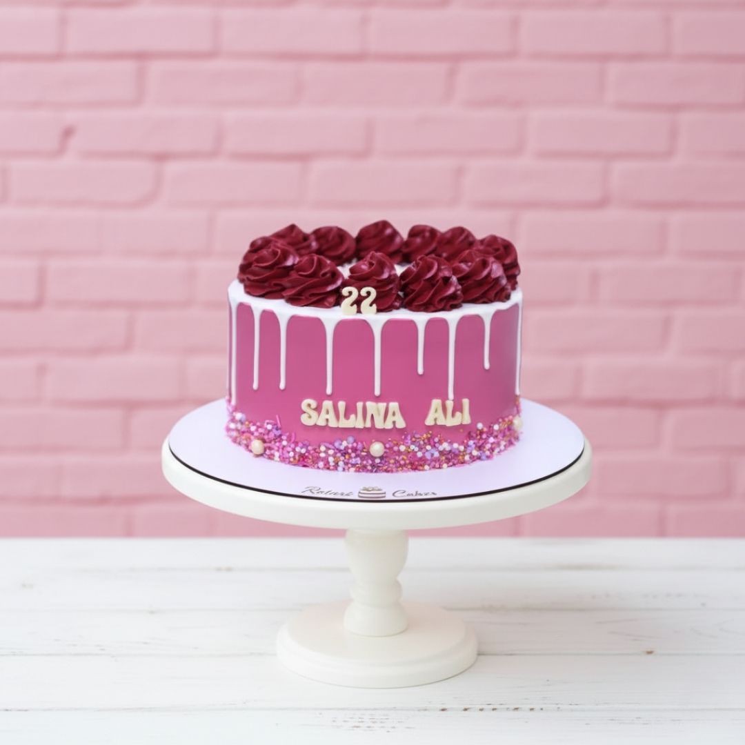 Pink cake with red roses and '22' on a white stand against a pink brick wall.