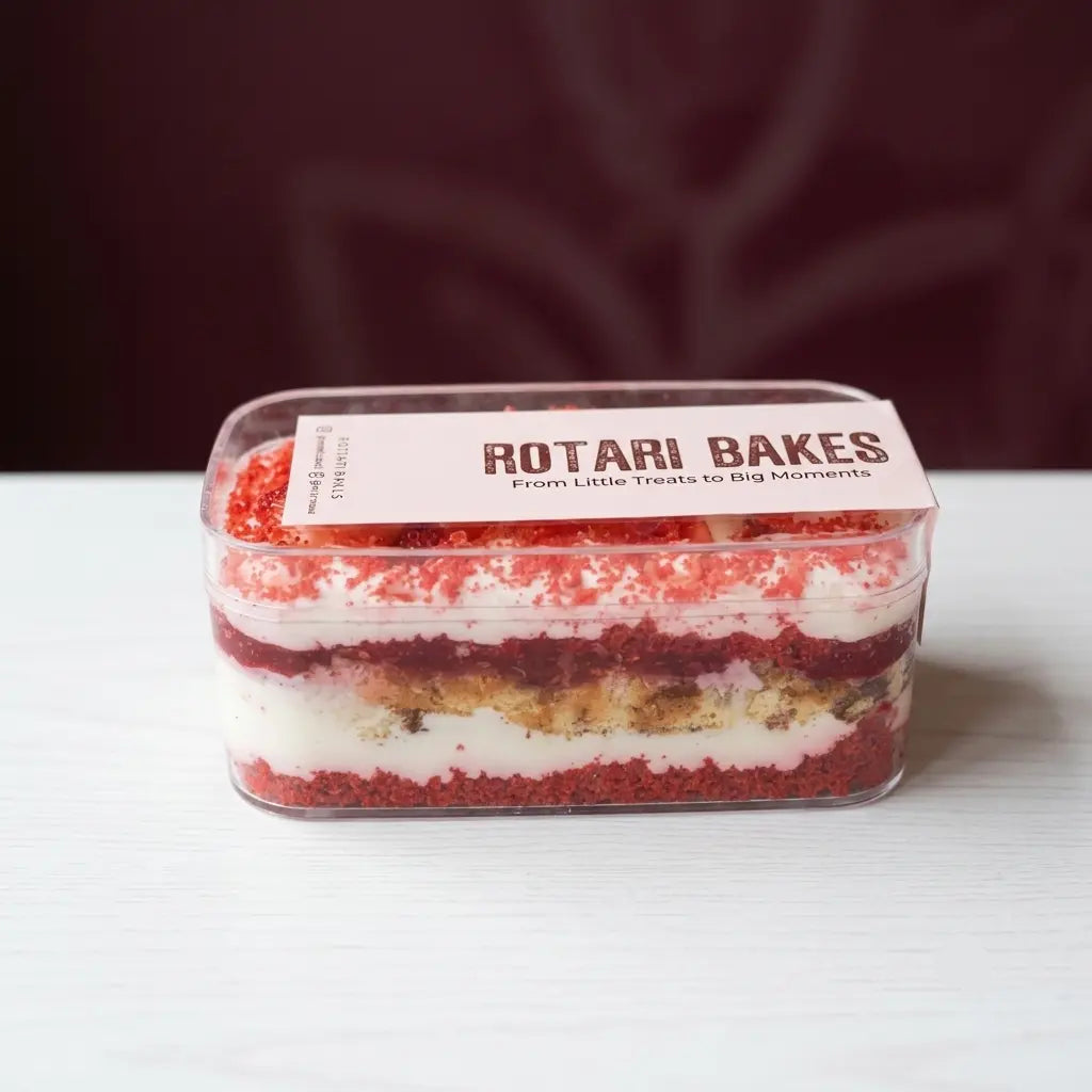A rectangular clear plastic container of red velvet cake layers with a white paper label reading 'ROTARI BAKES' on a white tabletop against a dark maroon background.