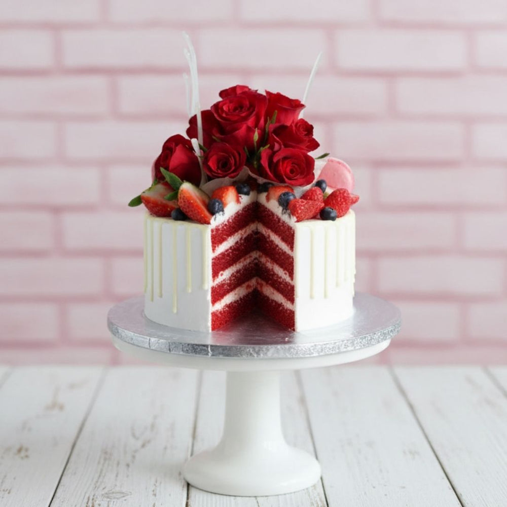 Red velvet cake with white frosting, berries, and red roses on a silver cake stand against a pink brick wall.
