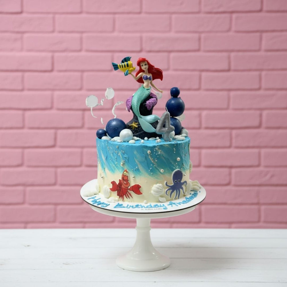 Underwater-themed birthday cake with mermaid and sea creature figurines against a pink brick wall.