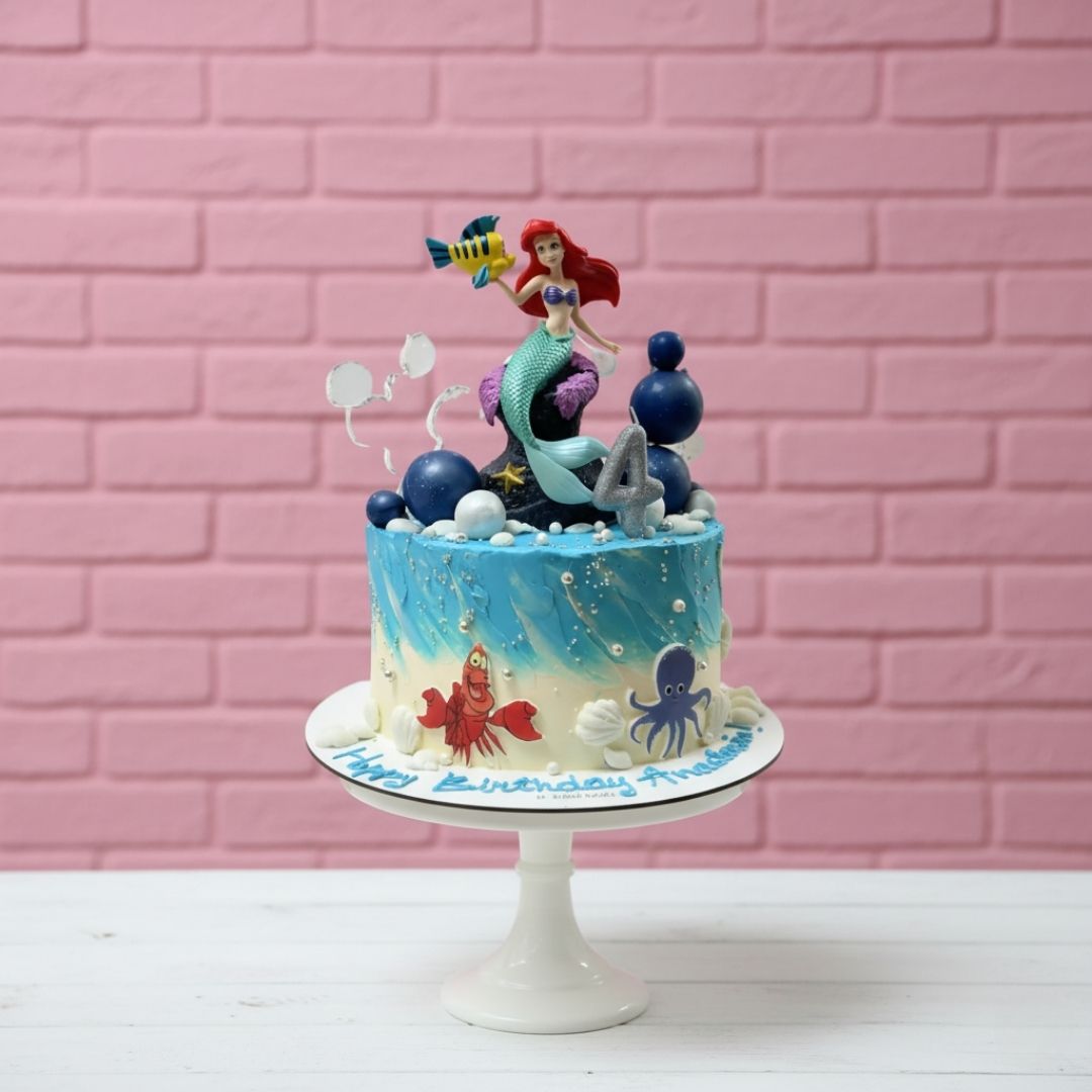 Underwater-themed birthday cake with mermaid and sea creature figurines against a pink brick wall.
