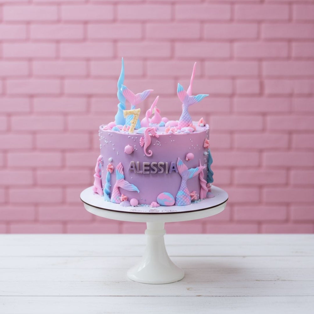 Pink birthday cake with mermaid-themed decorations on a white stand against a pink brick wall.