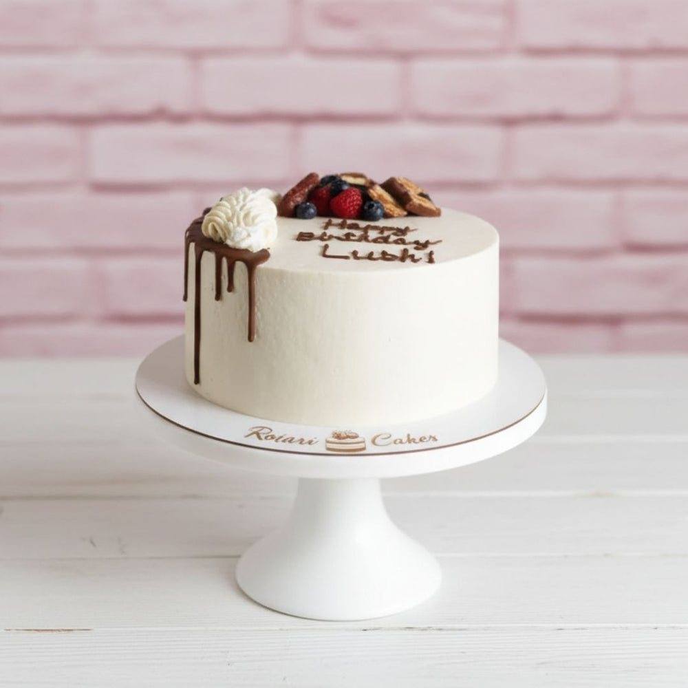 White birthday cake with chocolate drizzle and berries on a white stand against a pink brick wall.