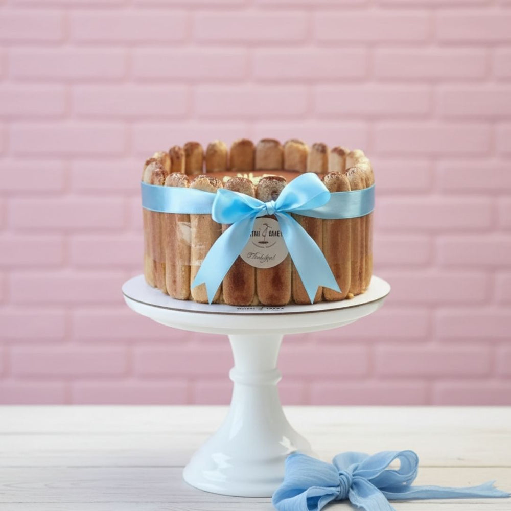tiramisu Cake with a blue ribbon on a white stand against a pink brick wall.