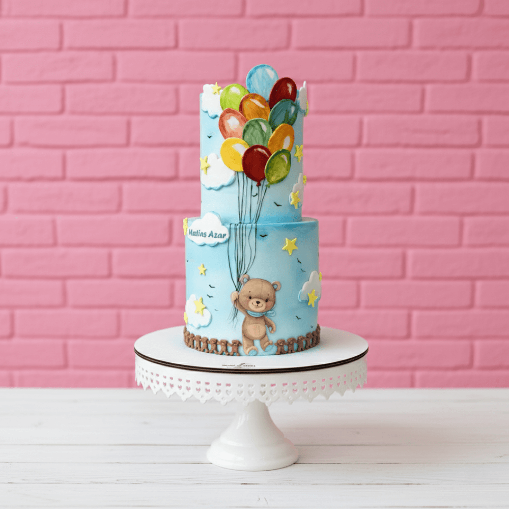 Two-tiered cake with teddy bear and balloons in blue colour against a pink brick wall.