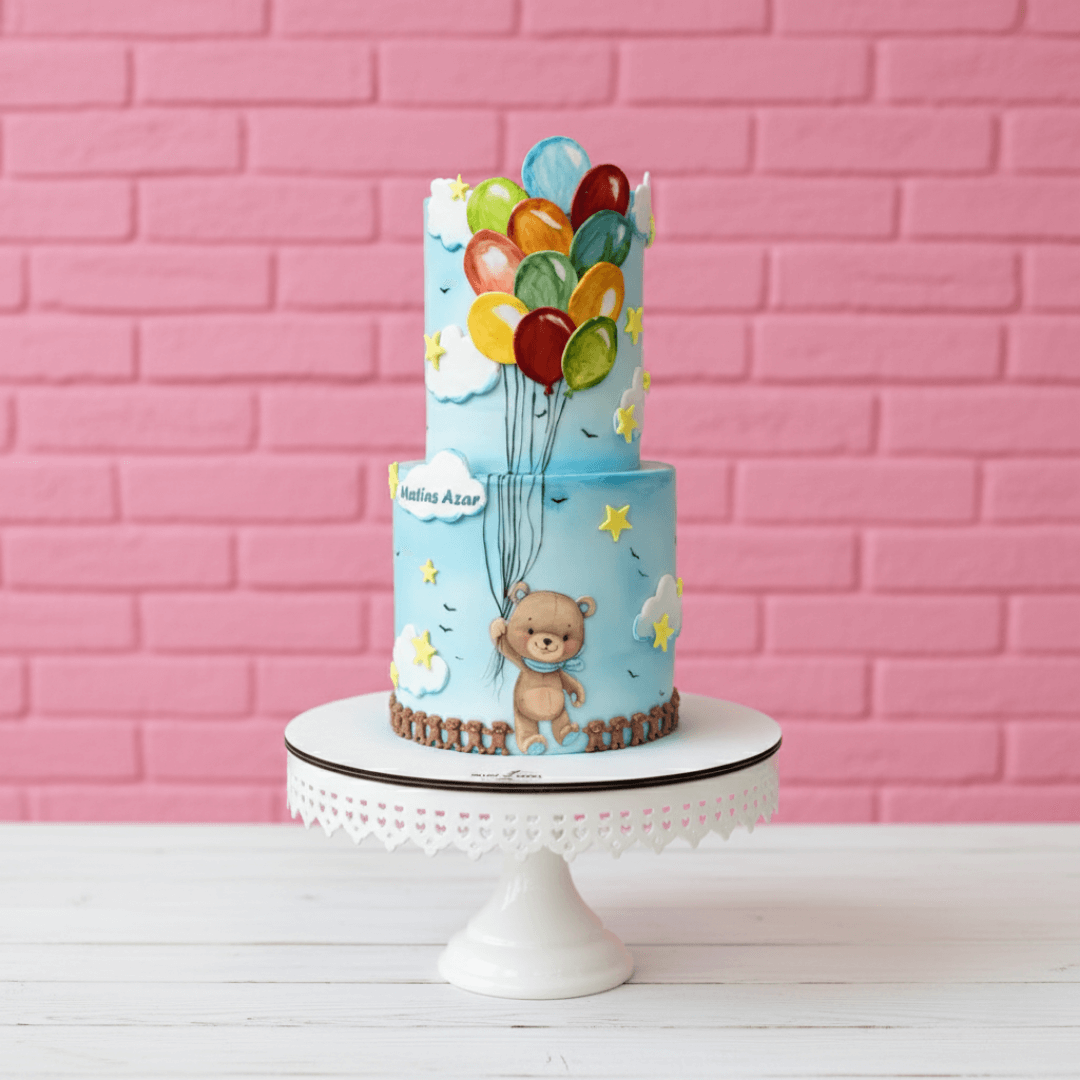 Two-tiered cake with teddy bear and balloons in blue colour against a pink brick wall.