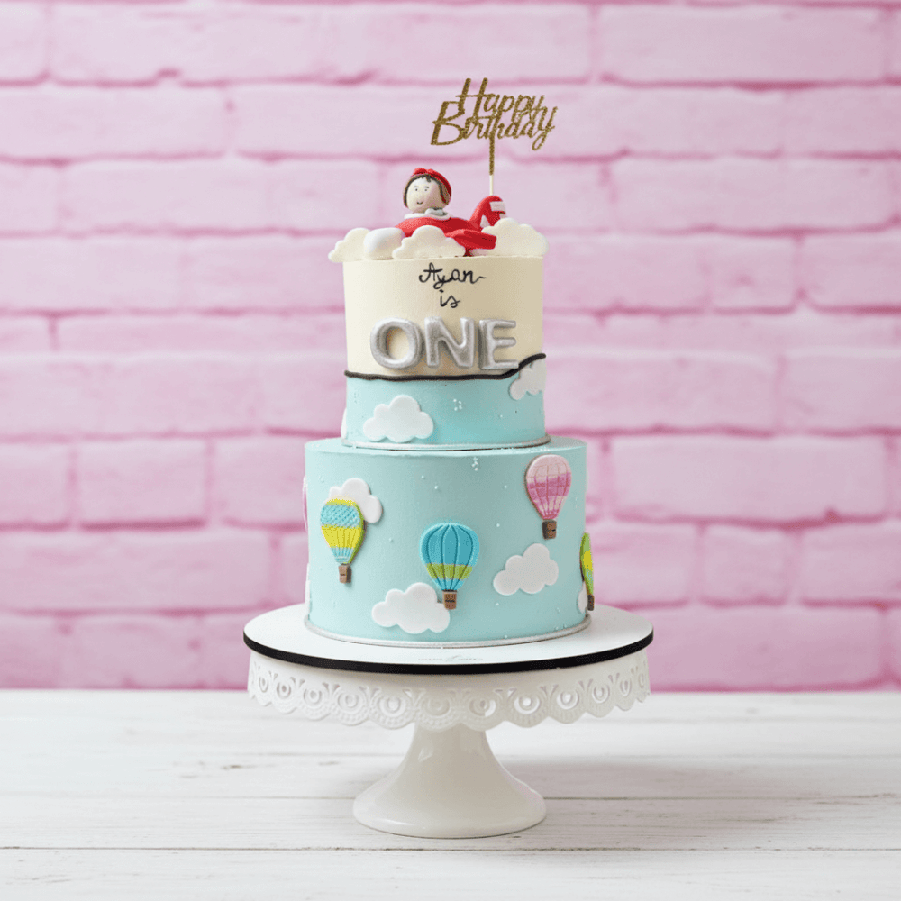 Two-tier birthday cake with hot air balloons and 'Happy Birthday' topper against a pink brick wall.