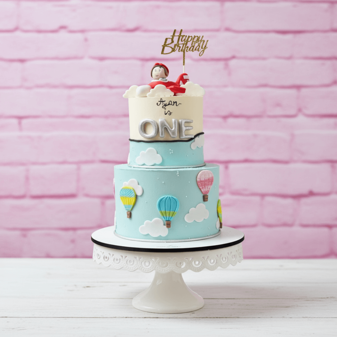 Two-tier birthday cake with hot air balloons and 'Happy Birthday' topper against a pink brick wall.
