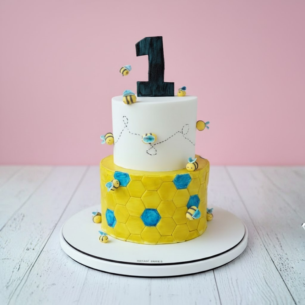 Two-tiered busy bee theme cake with yellow hexagonal pattern and white top tier, against a pink background
