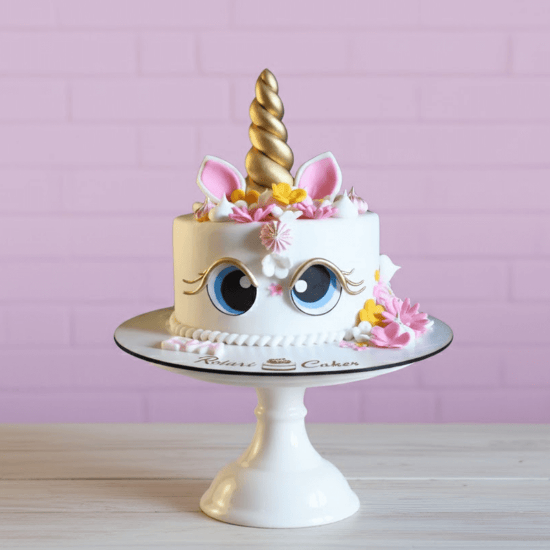 Unicorn cake with a gold horn and pink flowers on a white cake stand against a pink background