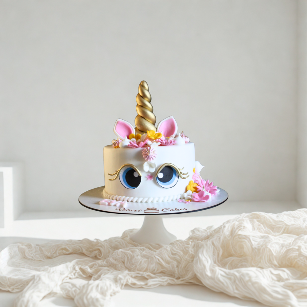 Unicorn cake with gold horn, pink ears, and blue eyes on a white background