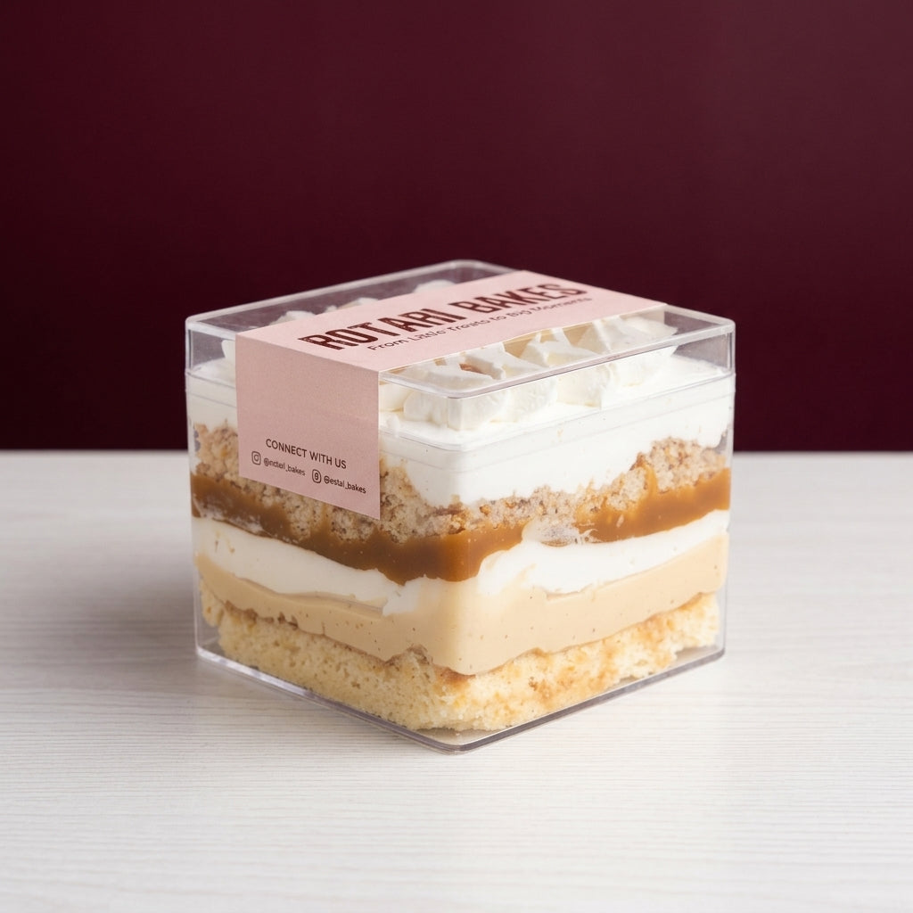 A square clear plastic container holding a layered cakes vanilla salted caramel and san sebastian cheesecake reading 'ROTARI BAKES' on a white tabletop against a dark burgundy background.