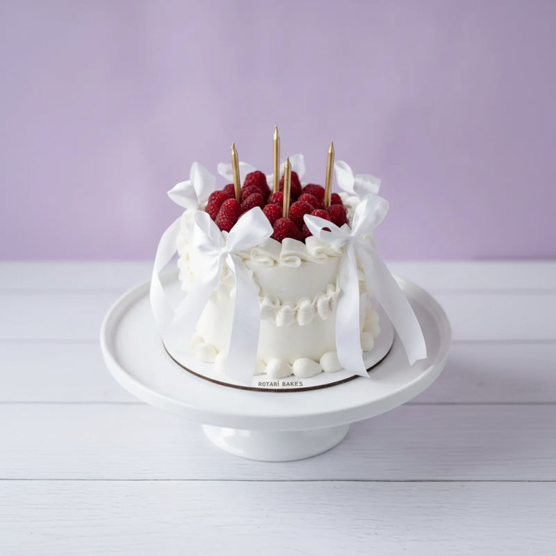 White cake with raspberries and gold candles on a white stand against a light purple background