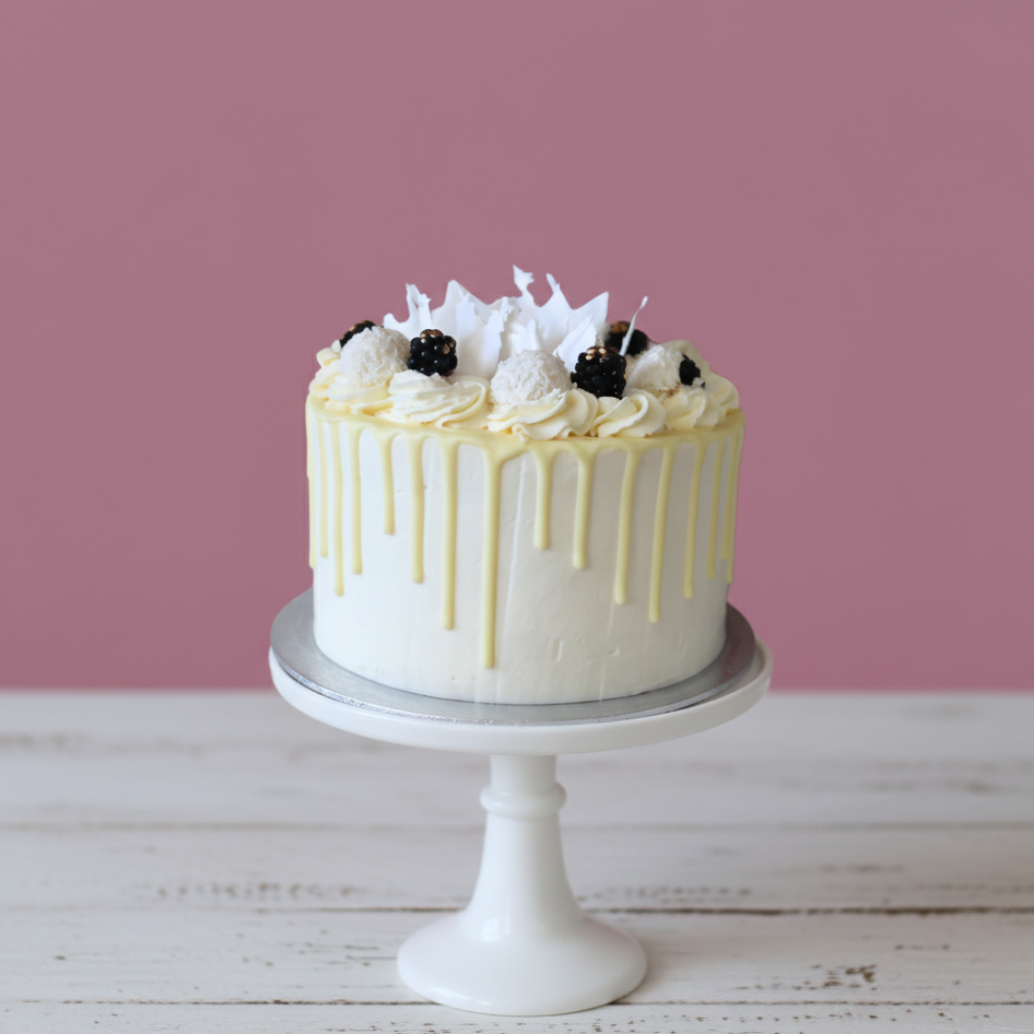 White cake with yellow drizzle and blackberries on a white stand against a pink background