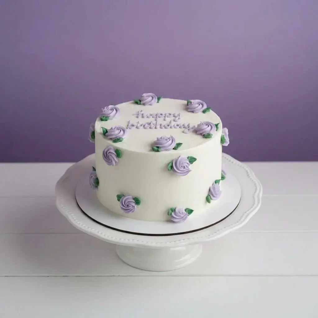 White birthday cake with purple flowers on a white stand against a purple background