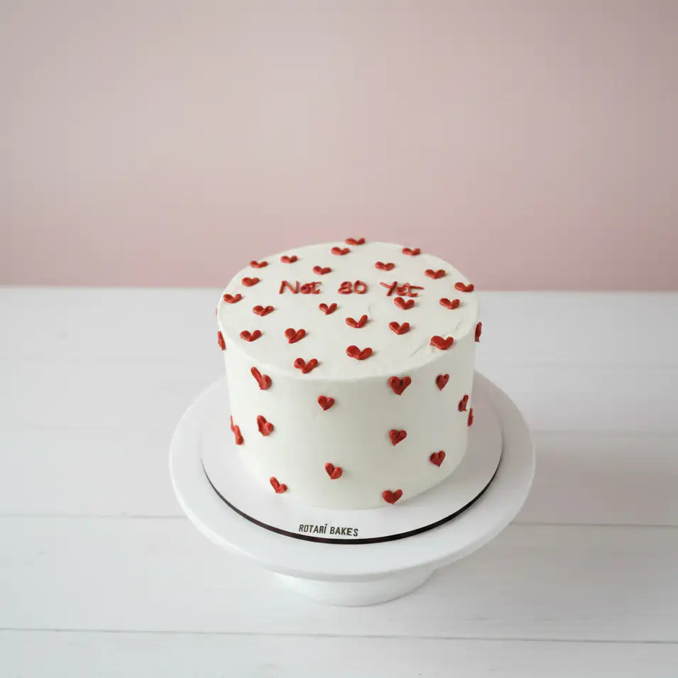 White mini cake with red heart decorations on a white stand against a light pink background