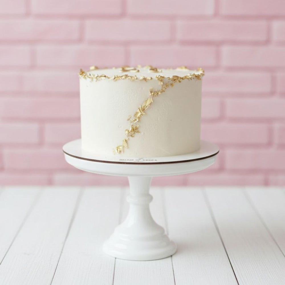 White cake with gold accents on a white stand against a pink brick wall.