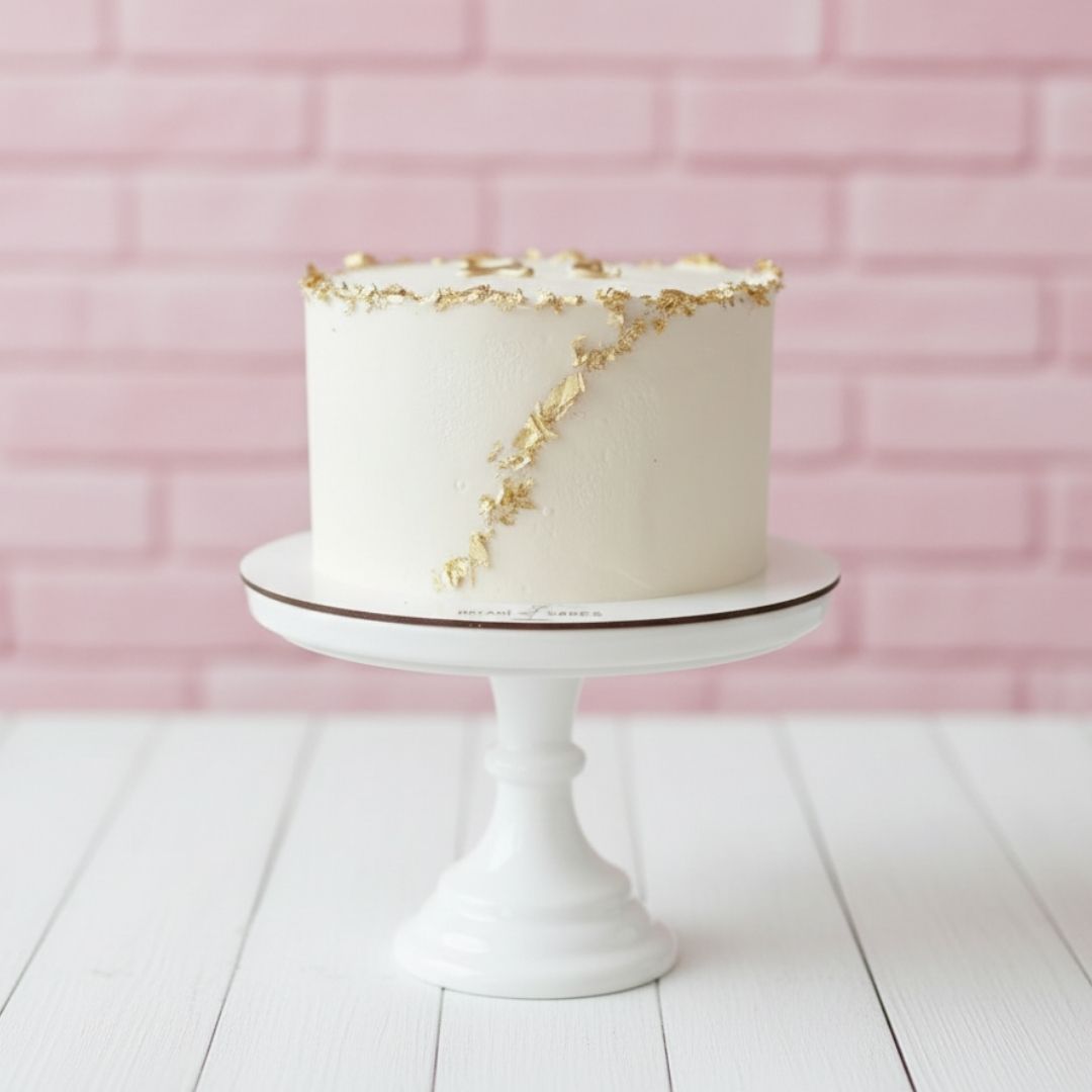 White cake with gold accents on a white stand against a pink brick wall.