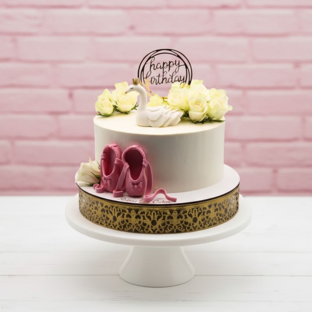 White birthday cake with pink ballet shoes, flowers, and 'happy birthday' topper on a pink brick background.